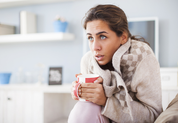 Woman sitting in her living room covered with blankets shivering and holding a red mug of a warm drink. 