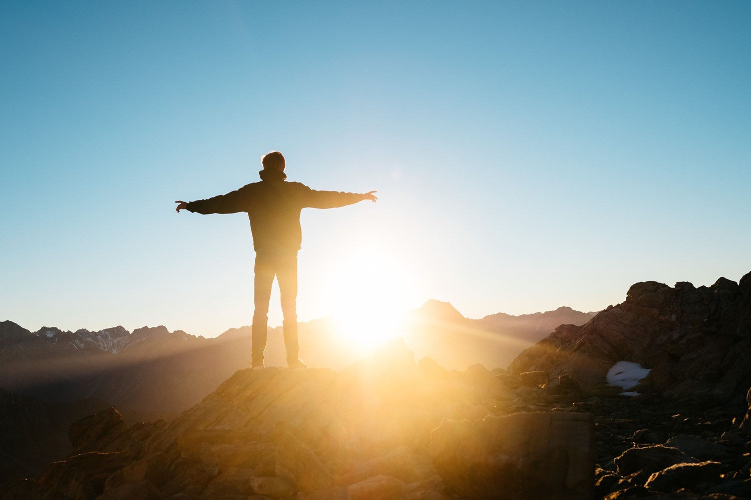 Man standing with his arms open onto of a mountain facing the sun that is setting. 