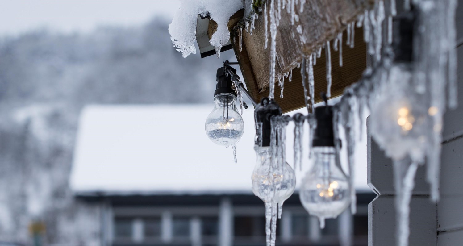 Icicles dripping down from a house covered in winter snow. Lights bulbs hang from the roof. 