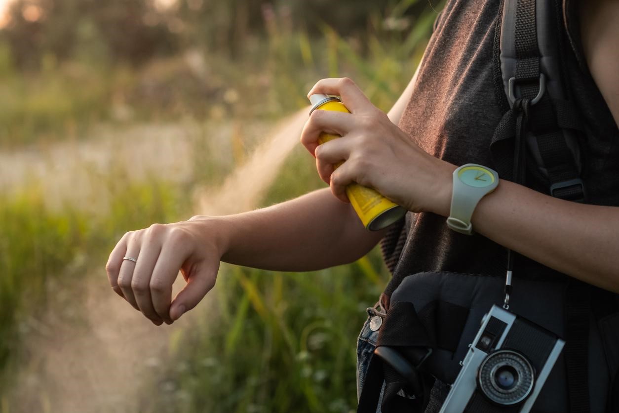 A woman in the outdoors spraying herself with bug spray to prevent mosquito bites. 